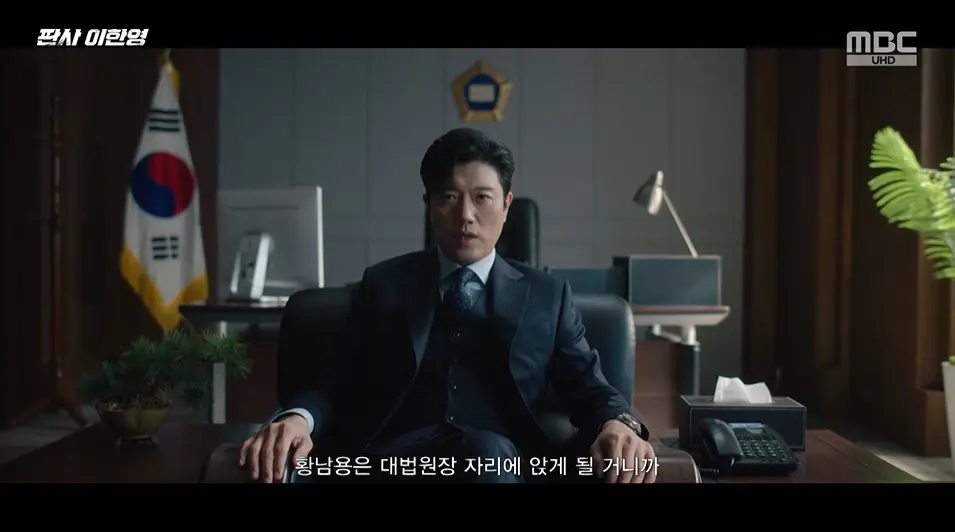 朴熙顺饰演的姜信镇在办公室观看新闻,对局势盲目自信